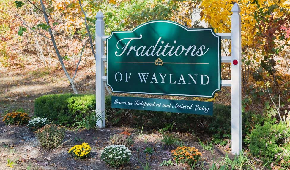 A decorative sign for Traditions of Wayland, surrounded by autumn foliage and vibrant flowers.