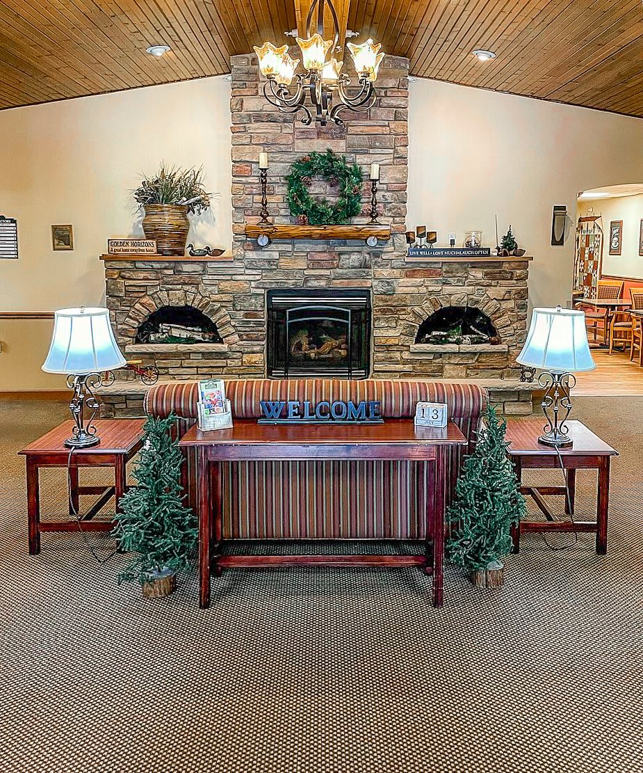 A cozy lobby area featuring a brick fireplace, decorated with a wreath and flanked by plants and lamps on wooden tables.