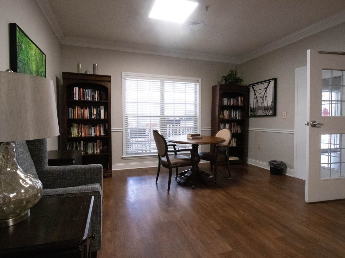 A cozy reading room with bookshelves, a table, and chairs by a large window.