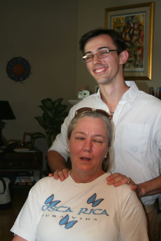 A smiling person standing behind another seated indoors in a relaxed setting.