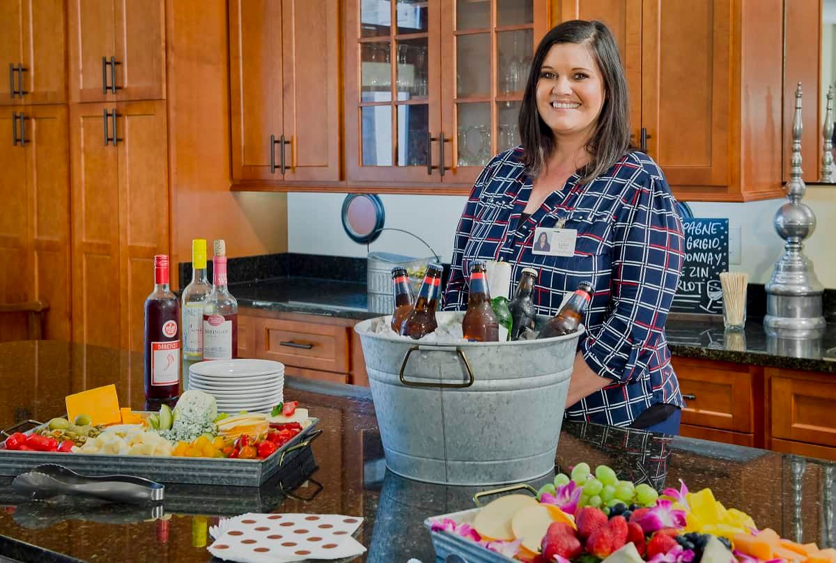 A woman stands in a kitchen with an assortment of drinks and snacks on the counter.