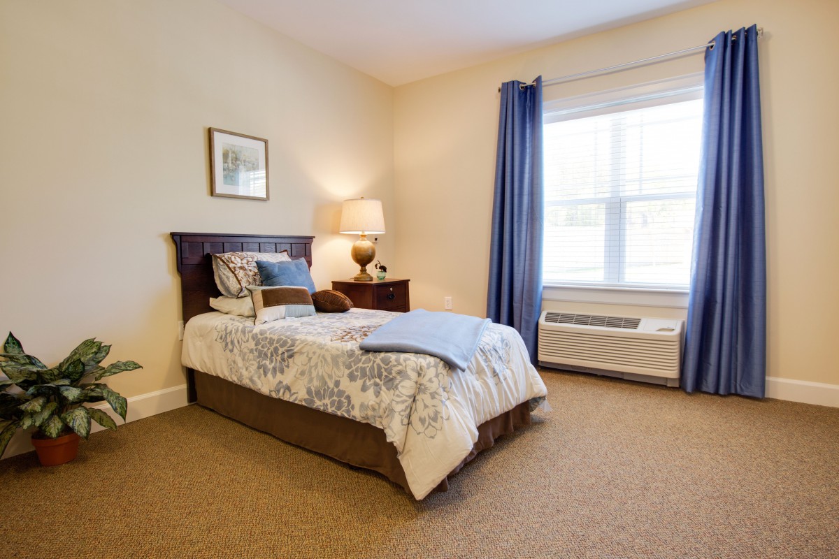 A cozy bedroom with a single bed, bedside table, lamp, and a large window with blue curtains.