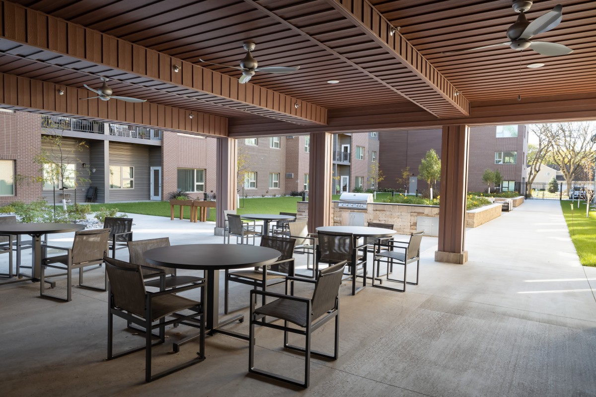 A covered patio area with tables and chairs overlooking a garden in an apartment complex.