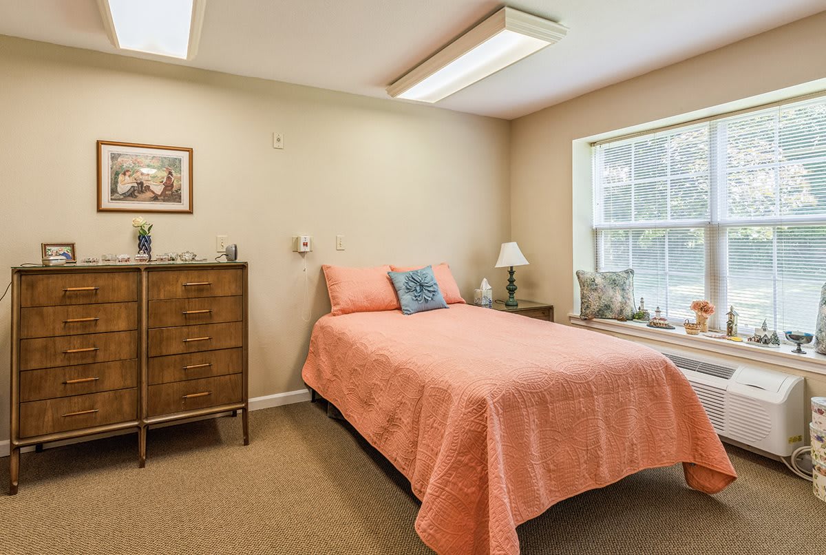 A cozy bedroom with a bed covered in a pink quilt, a dresser, and a lamp beside the window.