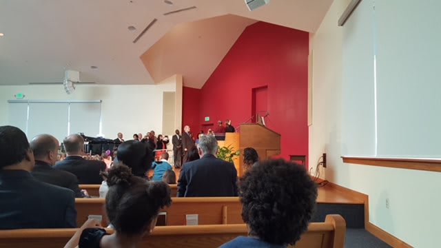 A church service in progress with congregation members seated and a speaker at the pulpit.