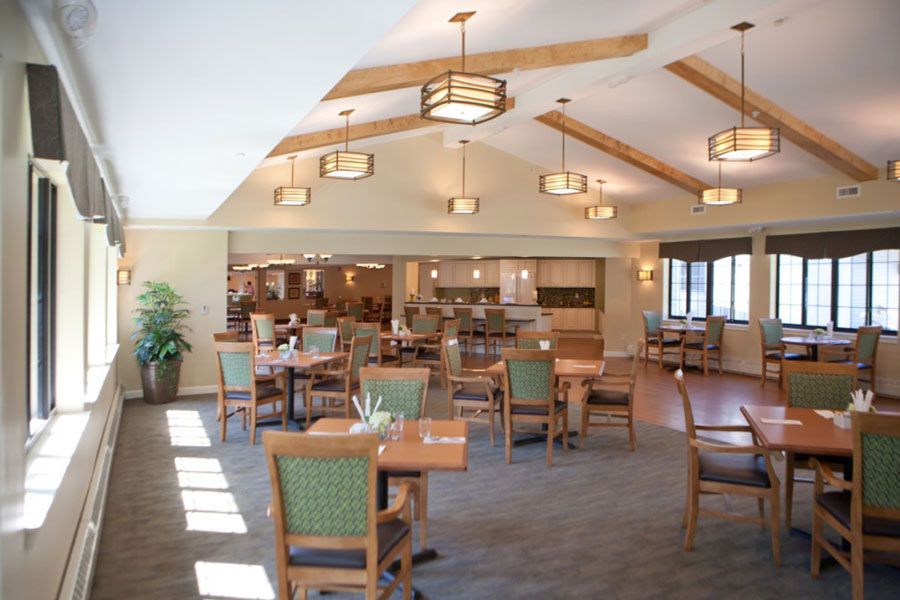A cozy dining room with arranged tables and chairs, featuring warm lighting and wooden ceiling beams.