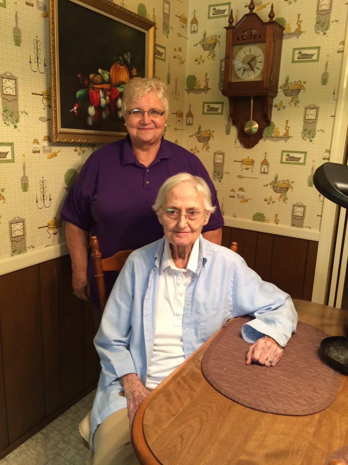 Two elderly individuals sitting and standing inside a room with decorated walls.