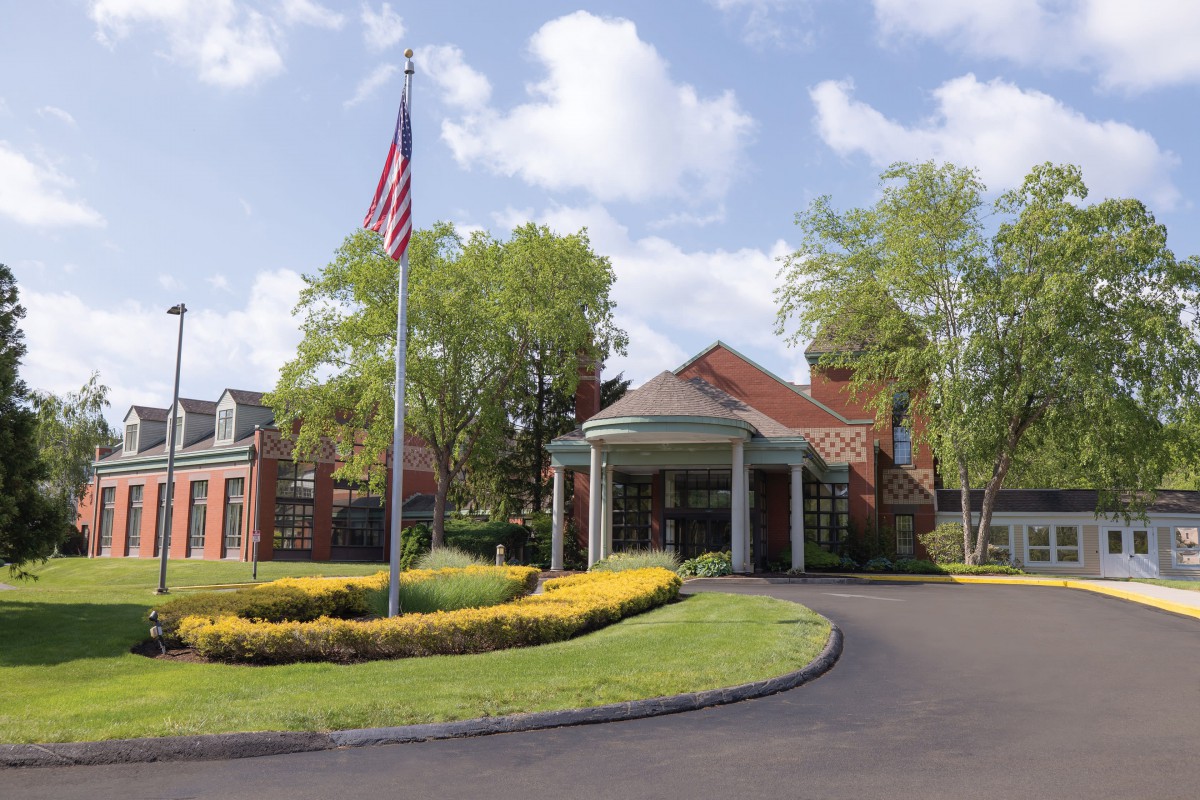 A brick building with a covered entrance, surrounded by lush landscaping and an American flag on a pole.