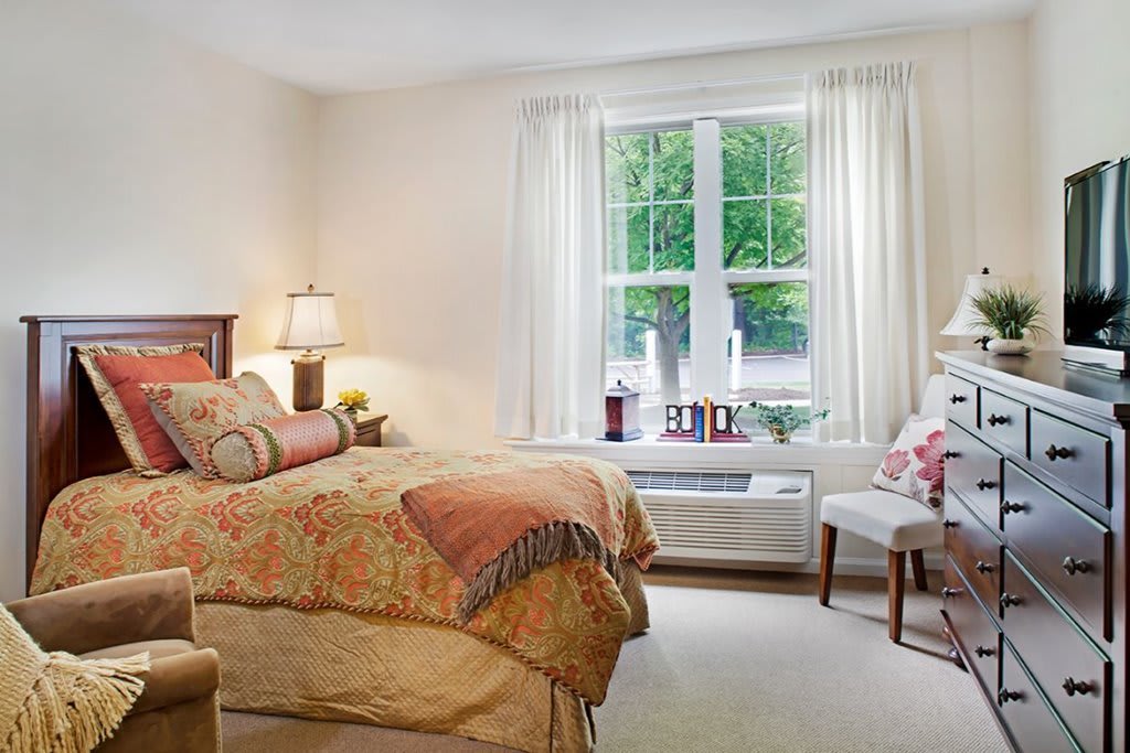 A cozy bedroom with a large window, featuring a bed with patterned bedding, a bedside lamp, and comfortable seating.