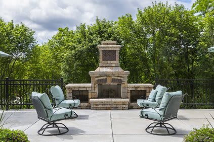 A stylish outdoor patio with a stone fireplace and cushioned chairs surrounded by lush greenery.