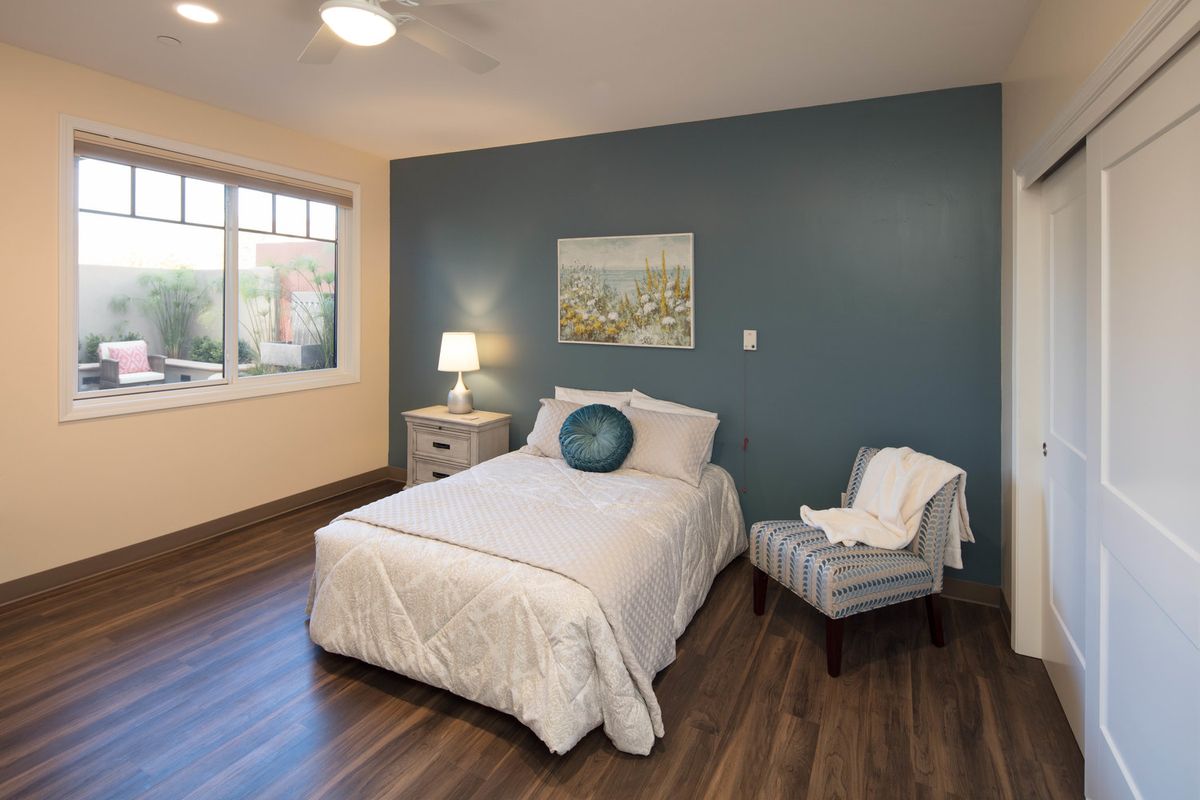 A cozy bedroom with a blue accent wall, featuring a bed, bedside lamp, chair, and large window.