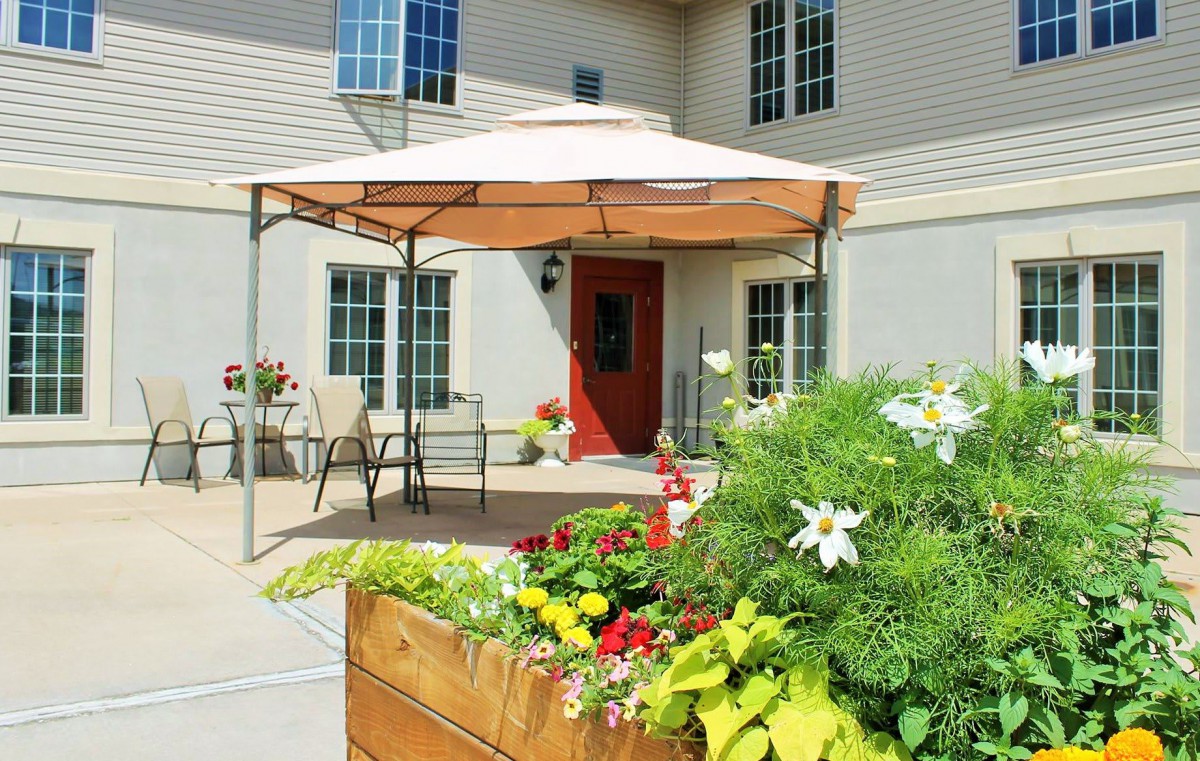 A cozy patio area with a gazebo and outdoor seating, surrounded by vibrant flowers and plants in front of a house with a red door.