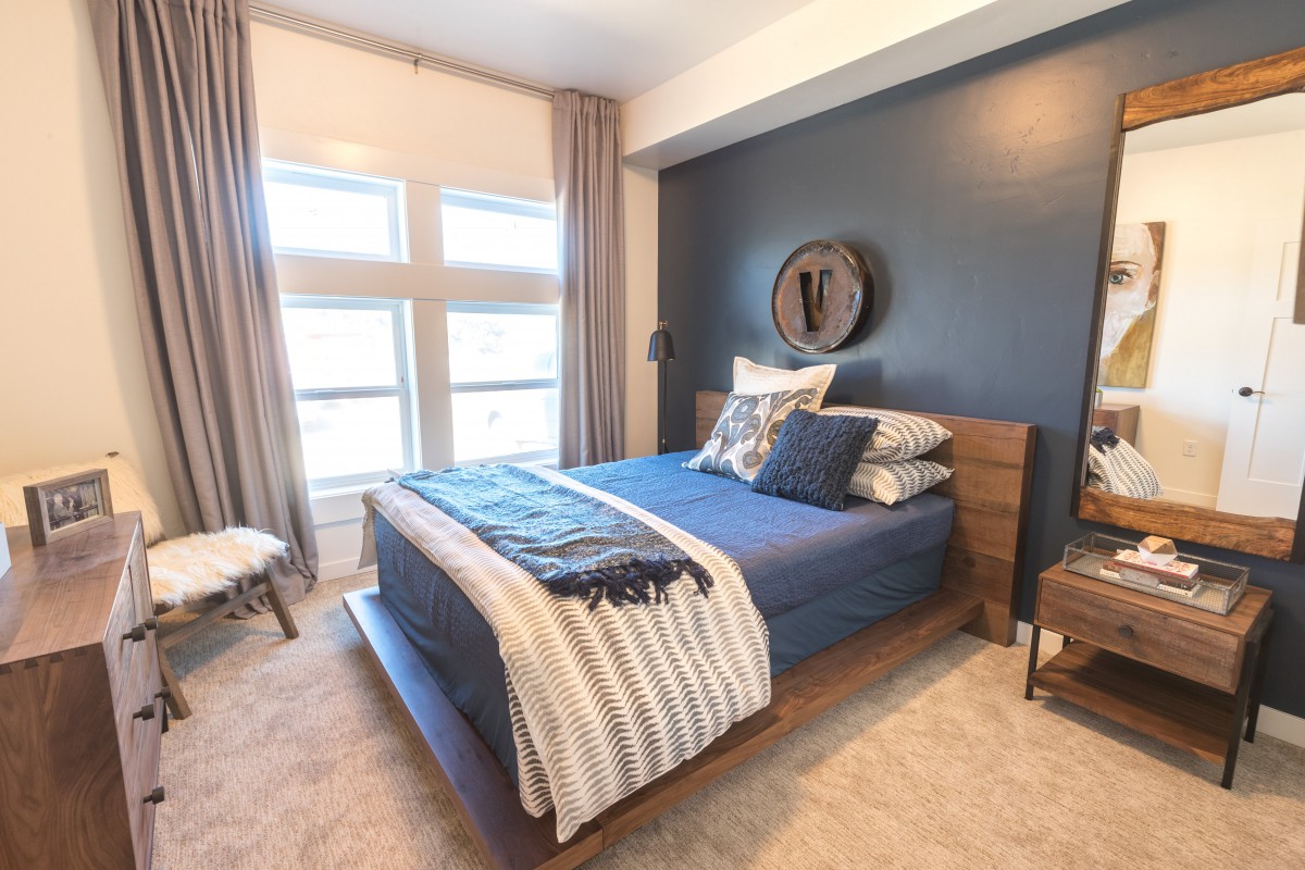 A modern bedroom with a blue accent wall and large windows, featuring a cozy bed adorned with pillows and blankets.