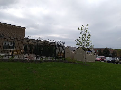 A view of a building surrounded by a fence, featuring a lone tree and grassy area under a cloudy sky.