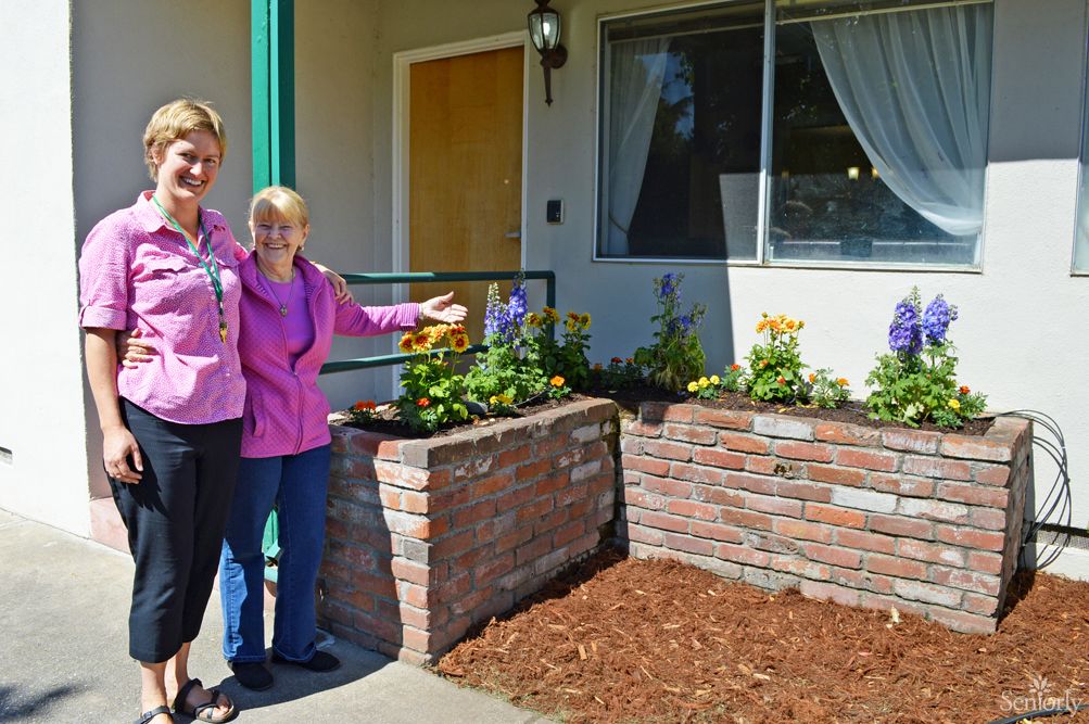 Two women proudly display a brick planter filled with colorful flowers outside a house.
