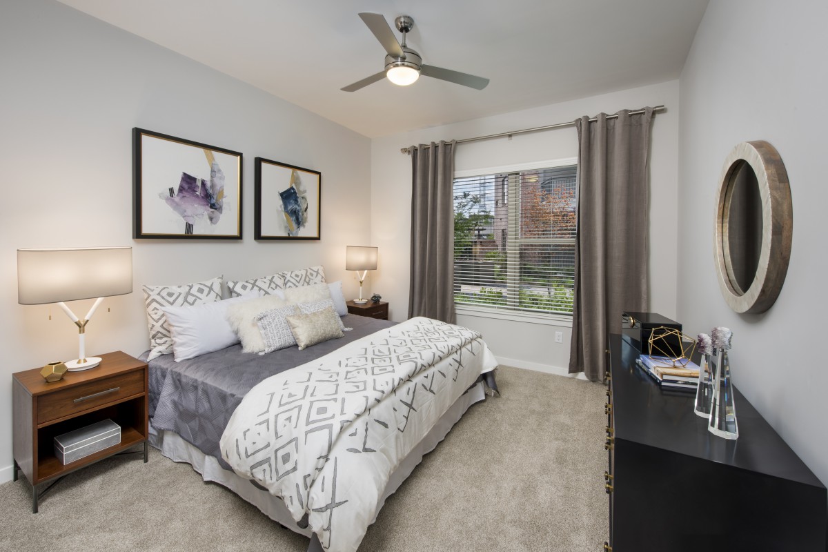 A modern bedroom with stylish decor, featuring a comfortable bed, ceiling fan, and large window with drapes.