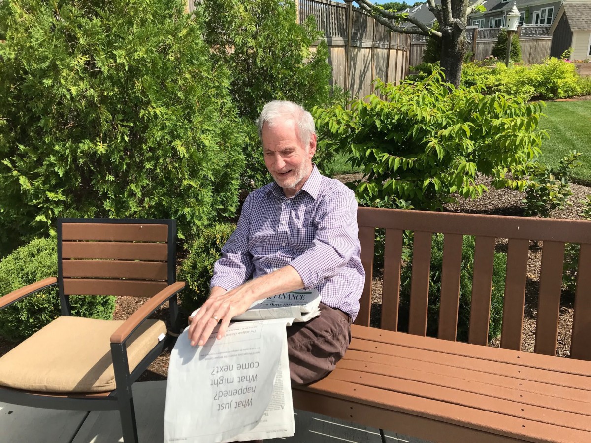 A man sitting on a bench in a garden, holding a book on a sunny day.