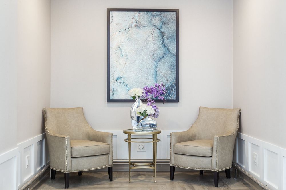 A cozy seating area with two beige chairs, a small round table holding a vase of flowers, and a large abstract painting on the wall.