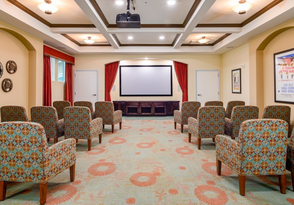 A cozy theater room with comfortable seating arranged in rows facing a large screen, accompanied by red curtains and a carpeted floor.