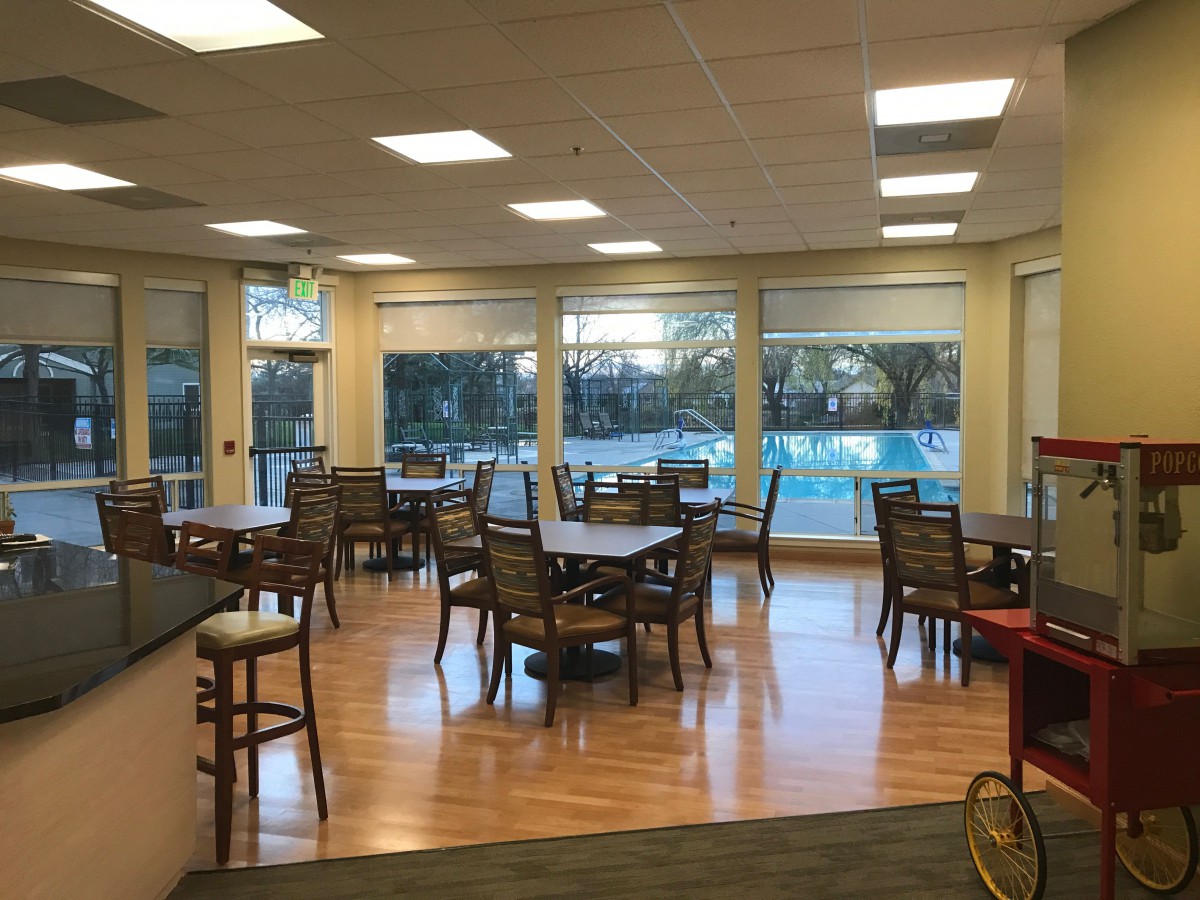 An empty dining area with tables and chairs, featuring large windows and wood flooring.