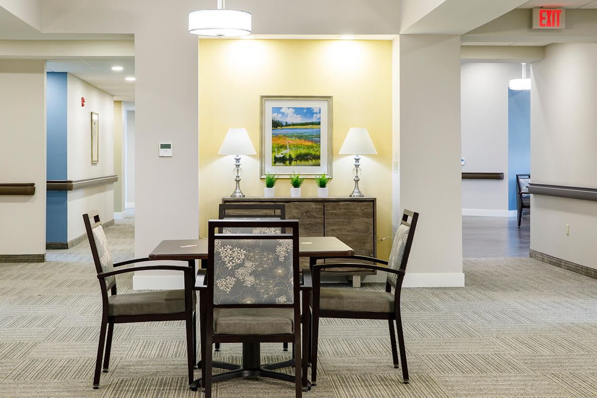 A well-lit dining area featuring a wooden table with four chairs, flanked by two lamps on a console table and a colorful landscape painting on the wall.