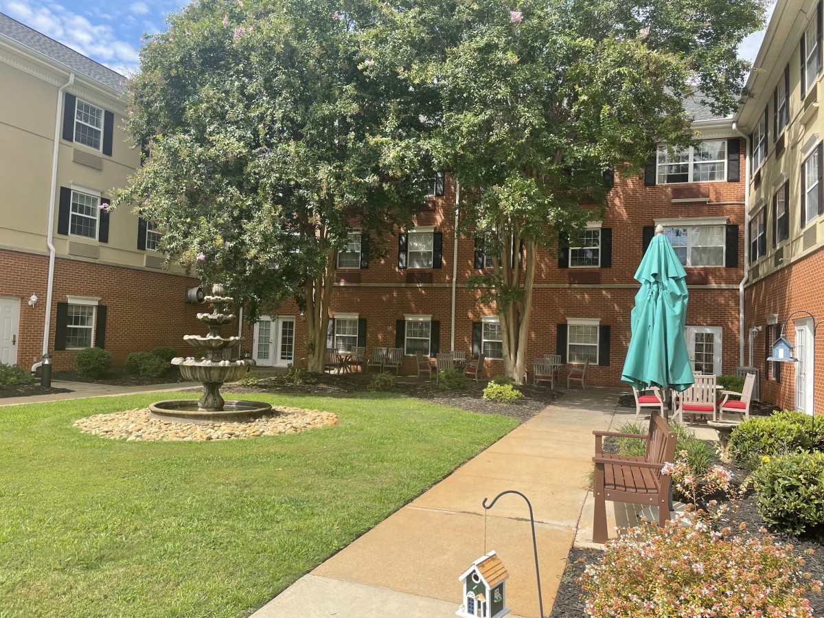 A peaceful garden area with a fountain, surrounded by trees and a multi-story building.