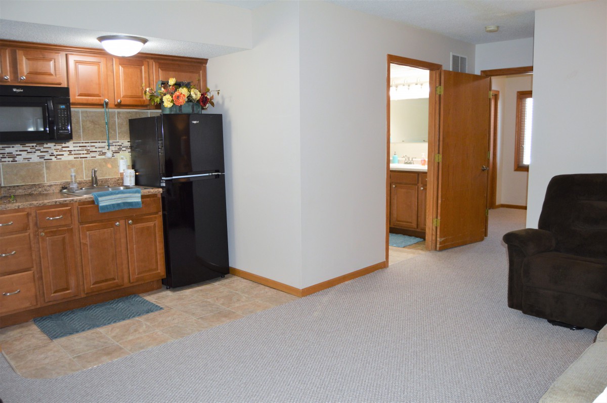 An interior view of an apartment featuring a small kitchen with wooden cabinets and a black fridge, alongside a carpeted hallway.