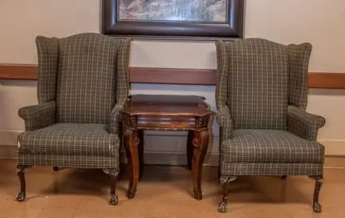 Two upholstered chairs flank a wooden side table, set against a wall with a framed painting.