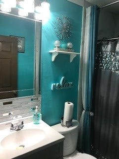 A stylish bathroom with turquoise walls, featuring a mirror above the sink, a decorative shelf with items, and a 'relax' sign.