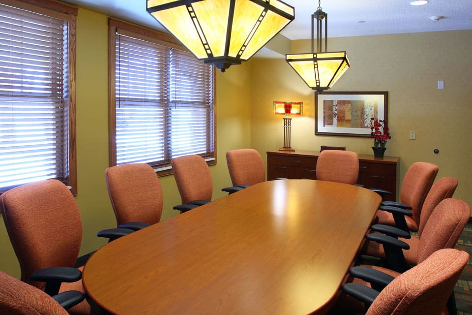 A modern conference room with a long wooden table, surrounded by several orange chairs and illuminated by stylish overhead lights.