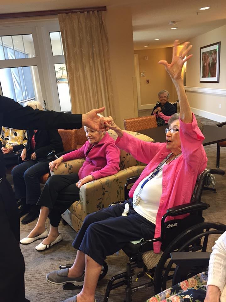 An elderly woman in a wheelchair reaching out during an interactive session at an assisted living facility.