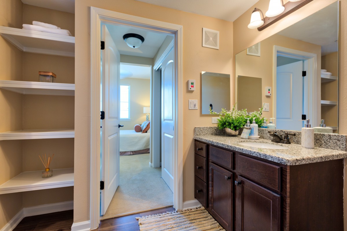 A modern bathroom with a vanity and mirror, leading to a cozy bedroom.