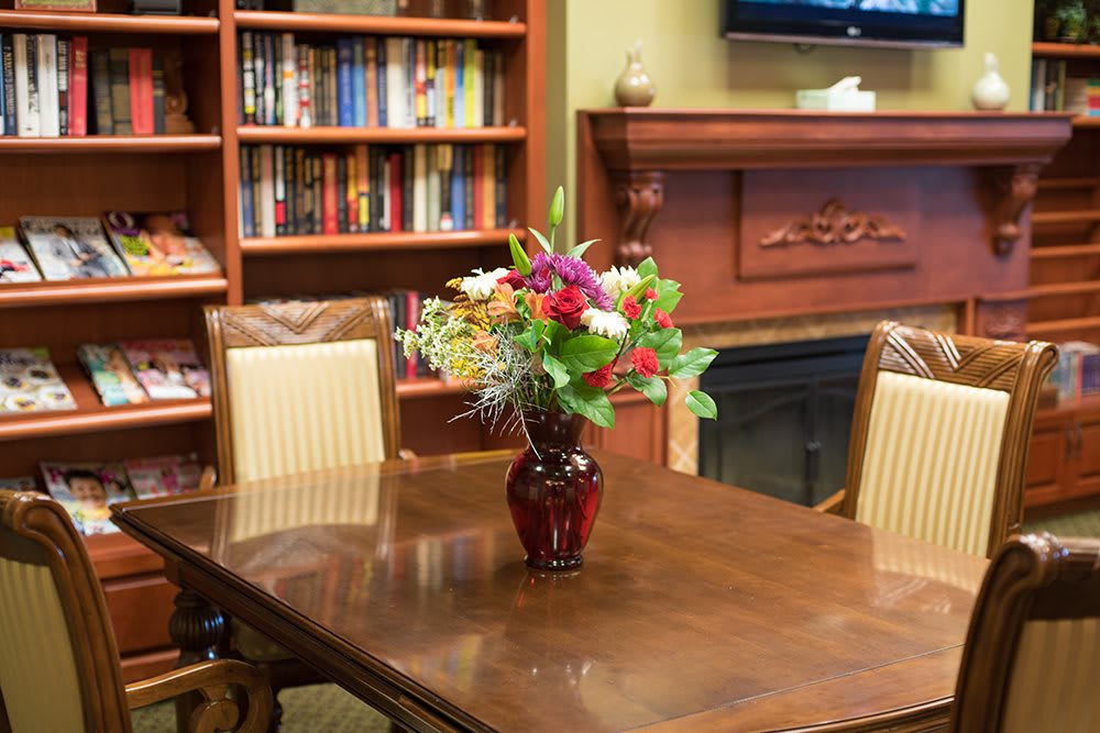 A cozy room with a wooden table adorned with a colorful flower arrangement in a vase, surrounded by bookshelves and a fireplace.