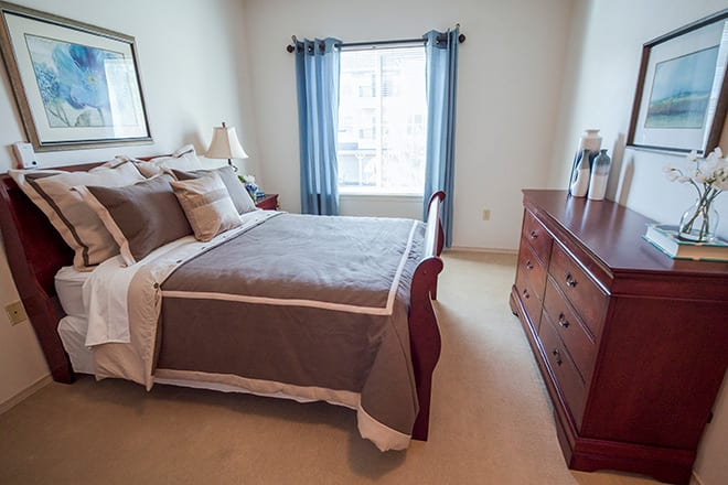A cozy bedroom with a neatly made bed, wooden dresser, and a window with blue curtains.