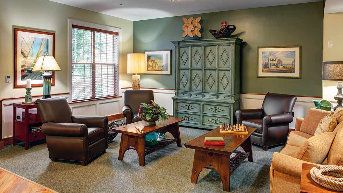 A cozy living room with comfortable armchairs, a wooden coffee table featuring a chessboard, and a large patterned cabinet against a green wall.