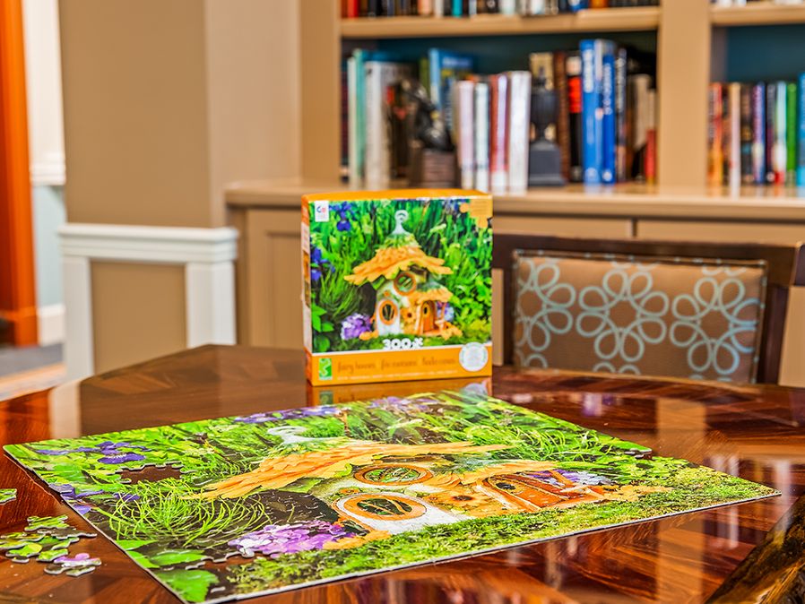 A completed jigsaw puzzle depicting a scenic fairy house, placed on a table with a bookshelf in the background.