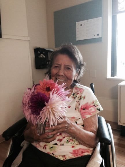 An elderly woman sitting in a wheelchair, smiling and holding a bouquet of flowers indoors.