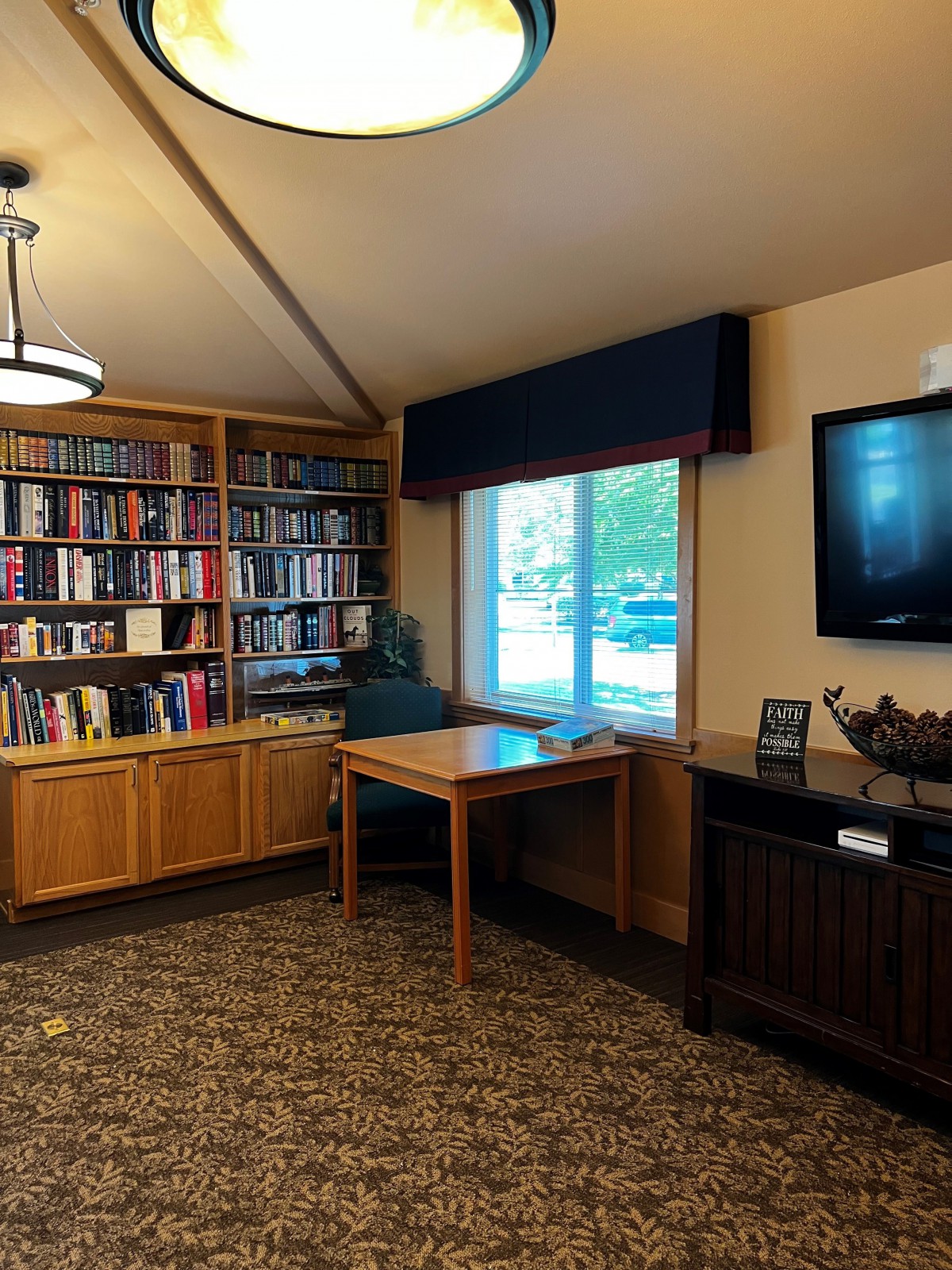 A cozy library corner with bookshelves, a table by the window, and a television on the wall.