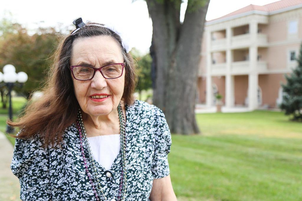 An elderly woman with glasses standing outside near a tree and building.