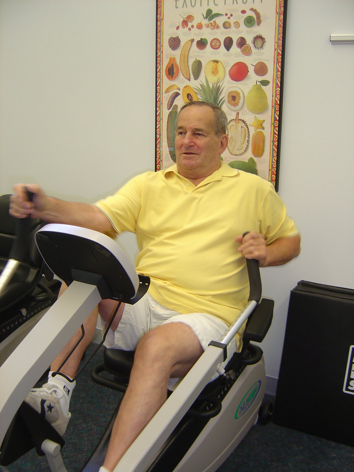A person exercising on a recumbent bike indoors, wearing a yellow shirt and white shorts.