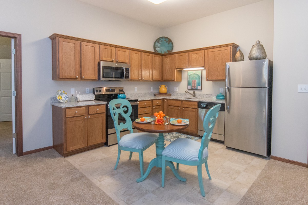A cozy kitchen with wooden cabinets and stainless steel appliances, featuring a small dining area with blue chairs and a round table.
