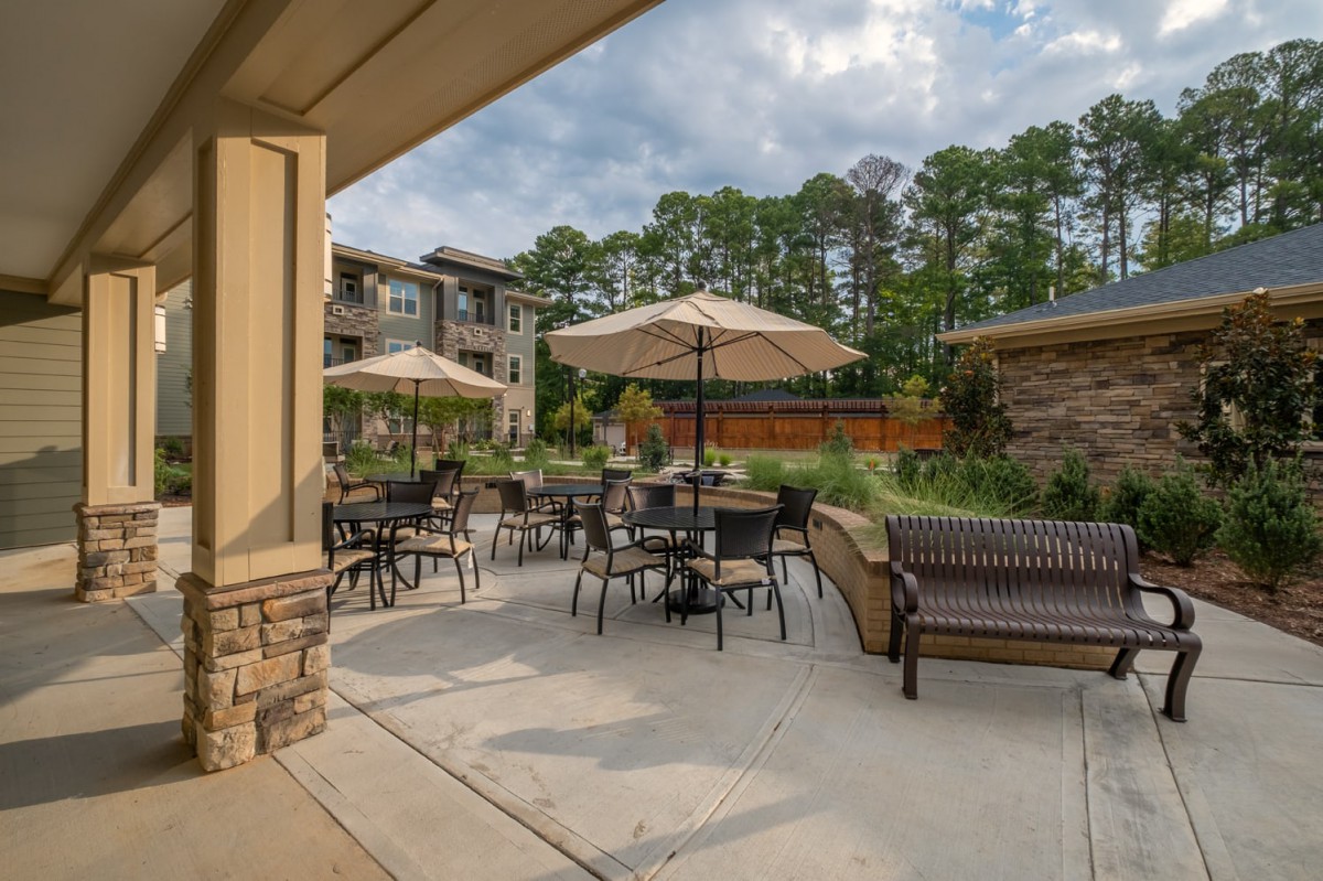 A peaceful outdoor patio with tables, chairs, and umbrellas set amidst an apartment complex with lush greenery.
