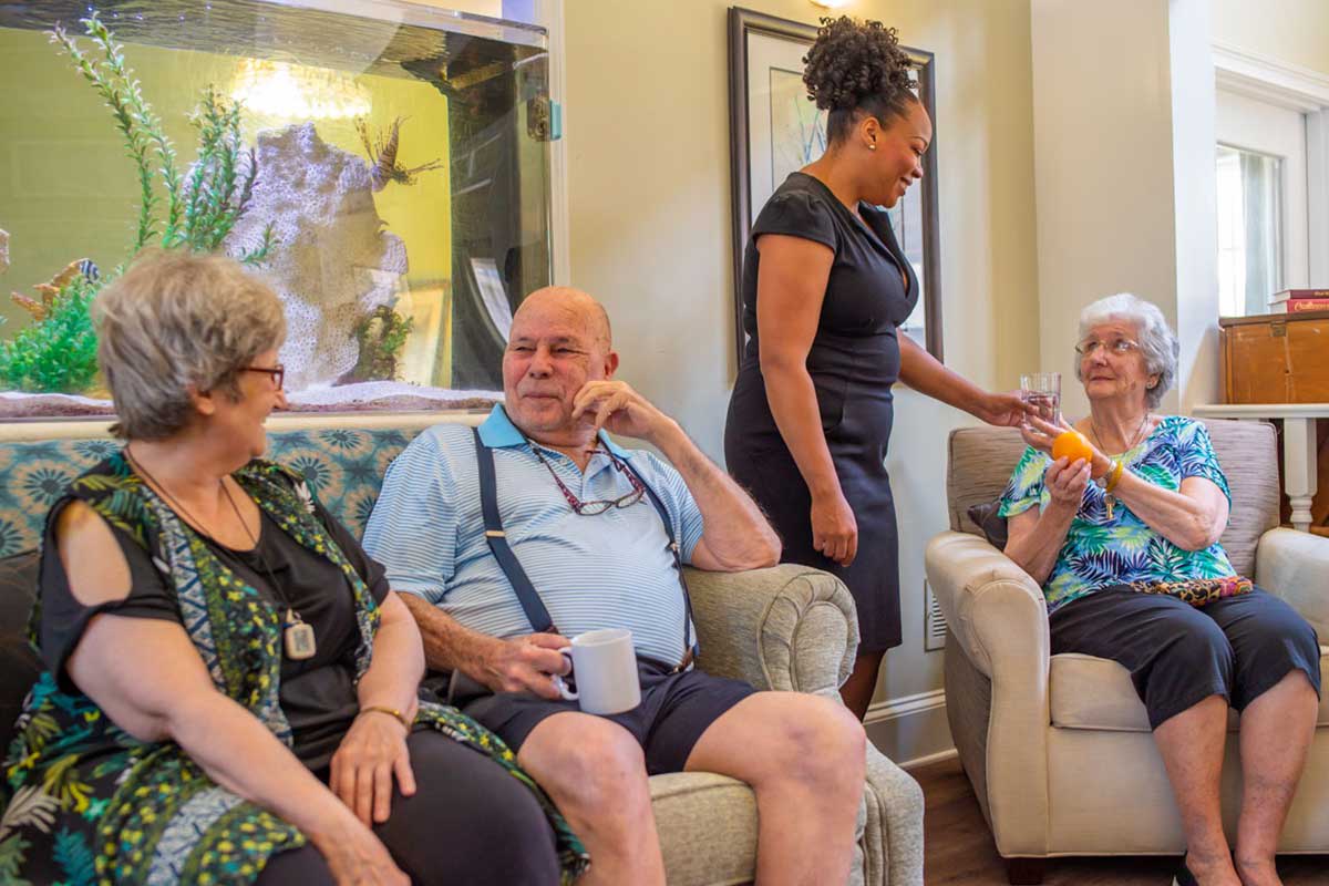 A group of seniors socializing in a living room, assisted by a caregiver.