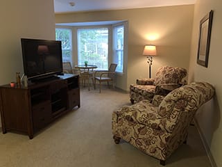 A cozy living room with patterned armchairs, a TV, and a warm lamp by a bay window.