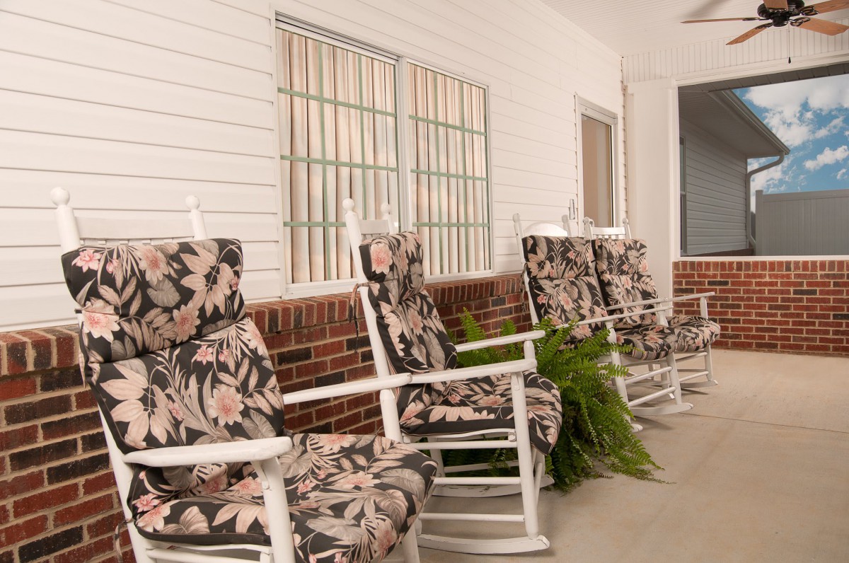 A row of floral-patterned rocking chairs line a porch with a brick wall and potted plants.