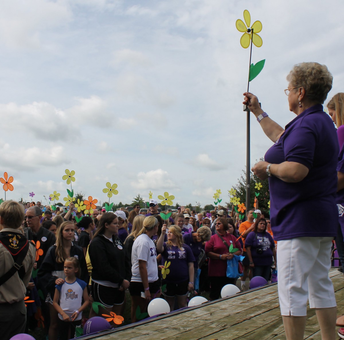 A large crowd gathers outdoors for a community event, with people holding colorful flower pinwheels.