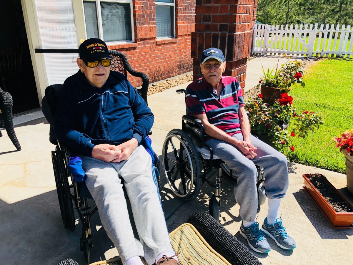 Two elderly individuals sitting in wheelchairs outside a brick building on a sunny day.