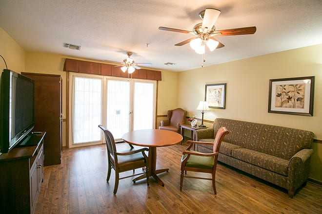 A cozy living room with a wooden floor, featuring a sofa, table, chairs, and ceiling fans.