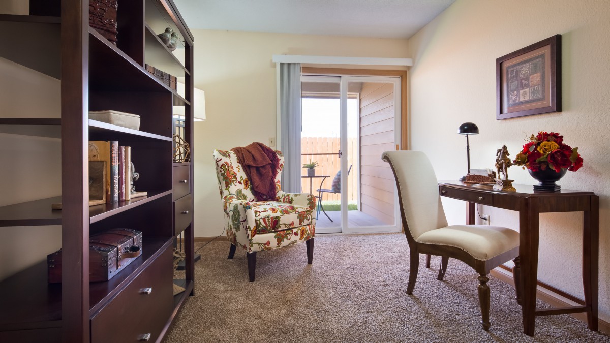 A cozy living room with an armchair, desk, and bookshelf, illuminated by natural light from the patio door.