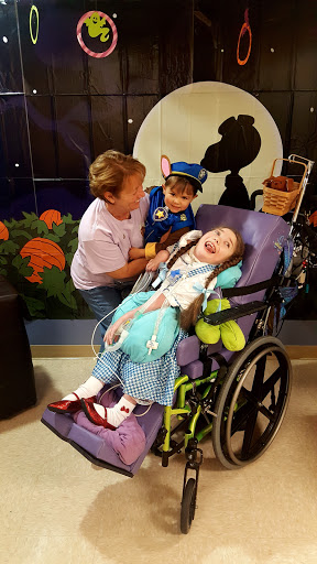 A young girl in a wheelchair enjoying time with her caregiver and a friendly costume character.